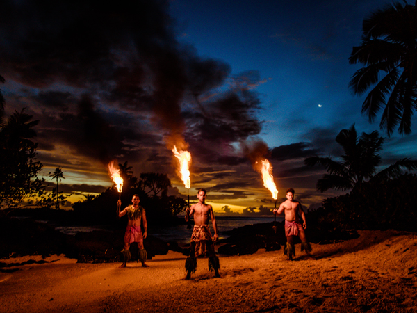 Three men in traditional dress stand on the beach at twilight, with sticks of fire