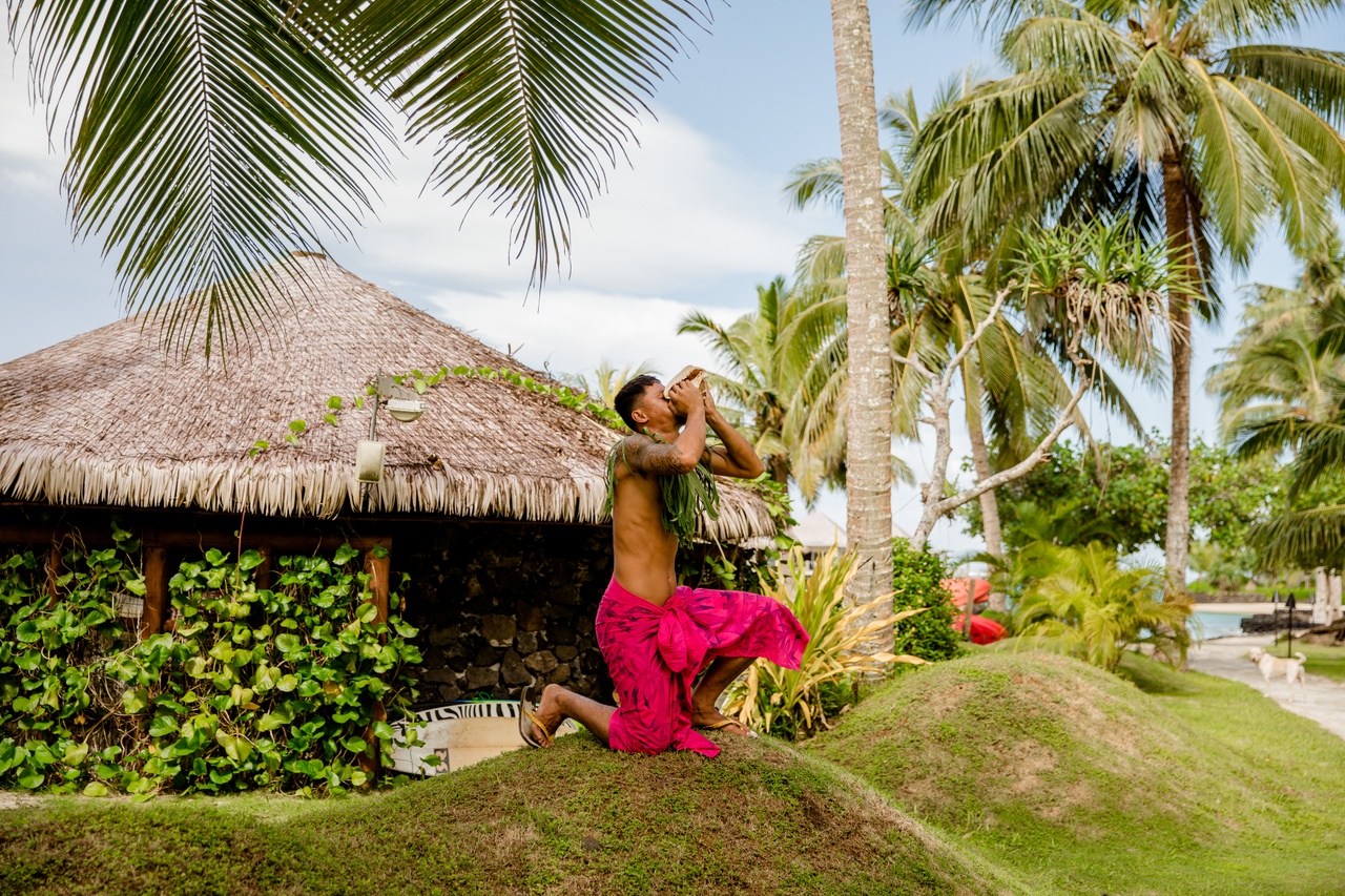 Samoan man blowing a conch shell beneath palm trees