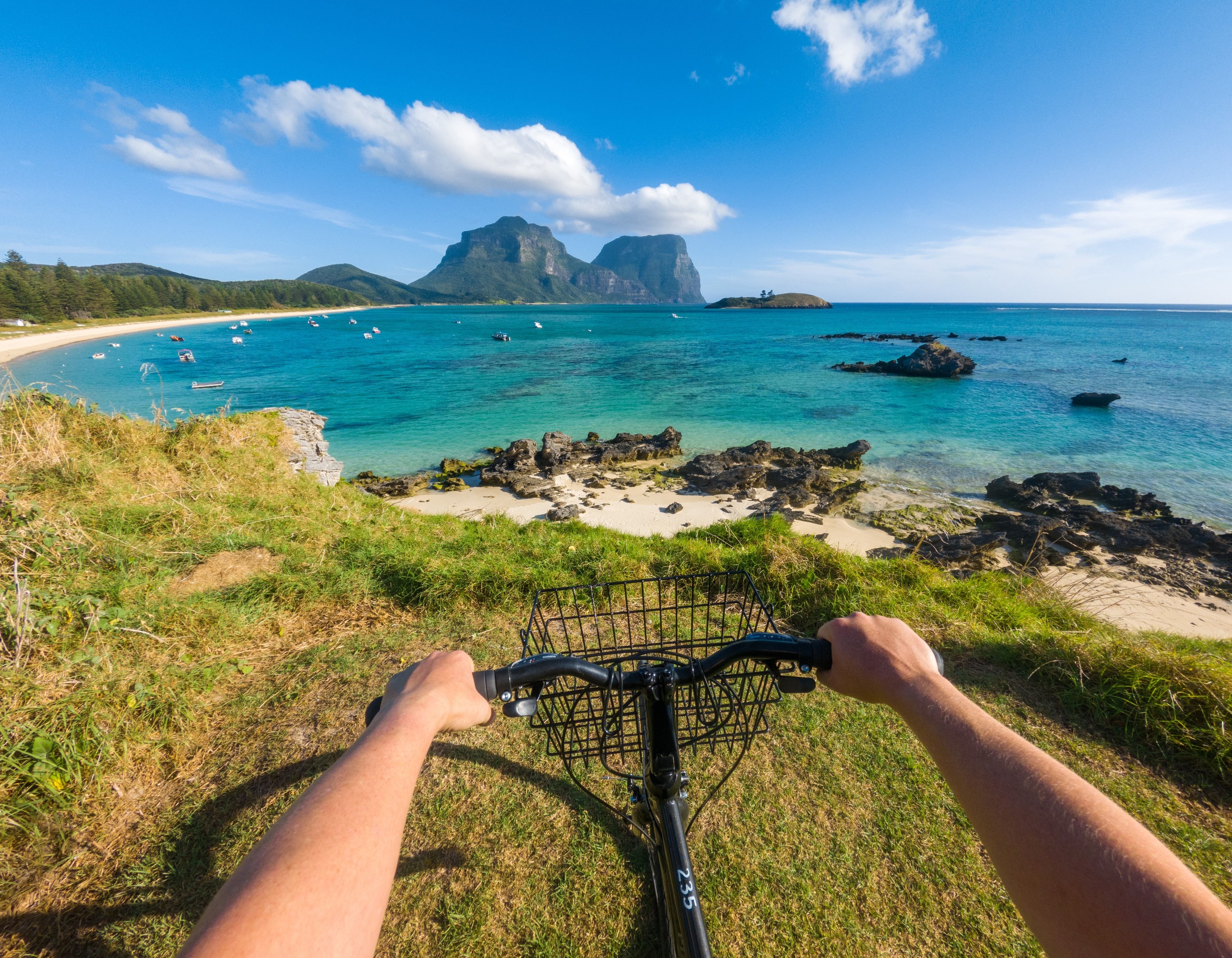 Hands appear on the handlebars of a bicycle, overlooking the sandy shoreline and distant mountains on Lord Howe Island