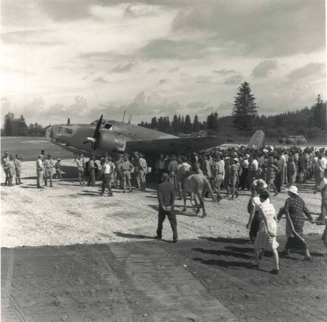 A black and white photo of a wartime airplane on the runway at Norfolk Island Airport, surrounded by locals