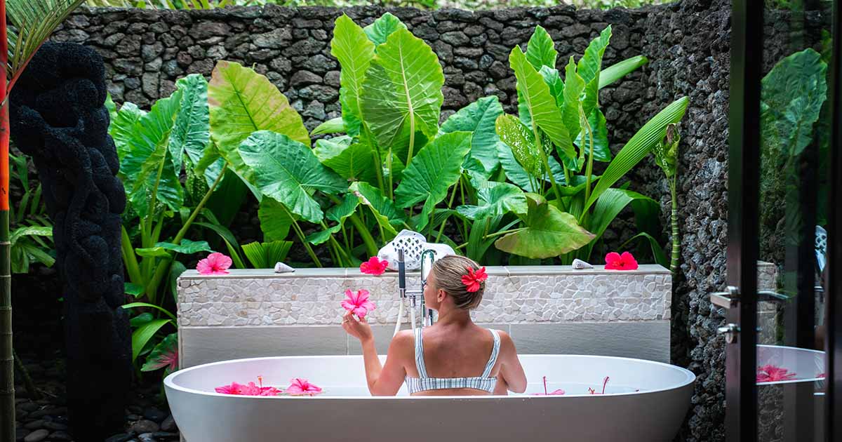 Replenish mind, body and soul with Health & Wellness experiences in Fiji A woman holds a flower while sitting in a white bath tub, surrounded by lush foliage and walls made from river stones.