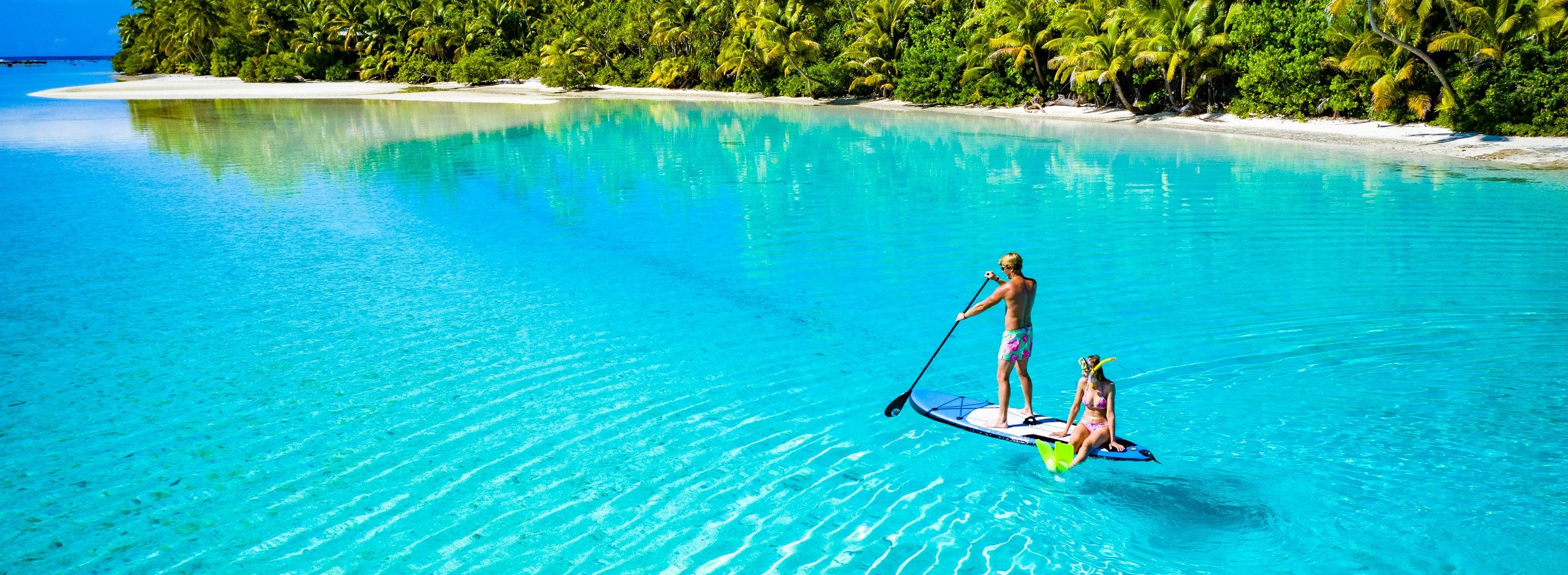 Woman in snorkelling gear sitting on back of paddleboard with man standing and paddling